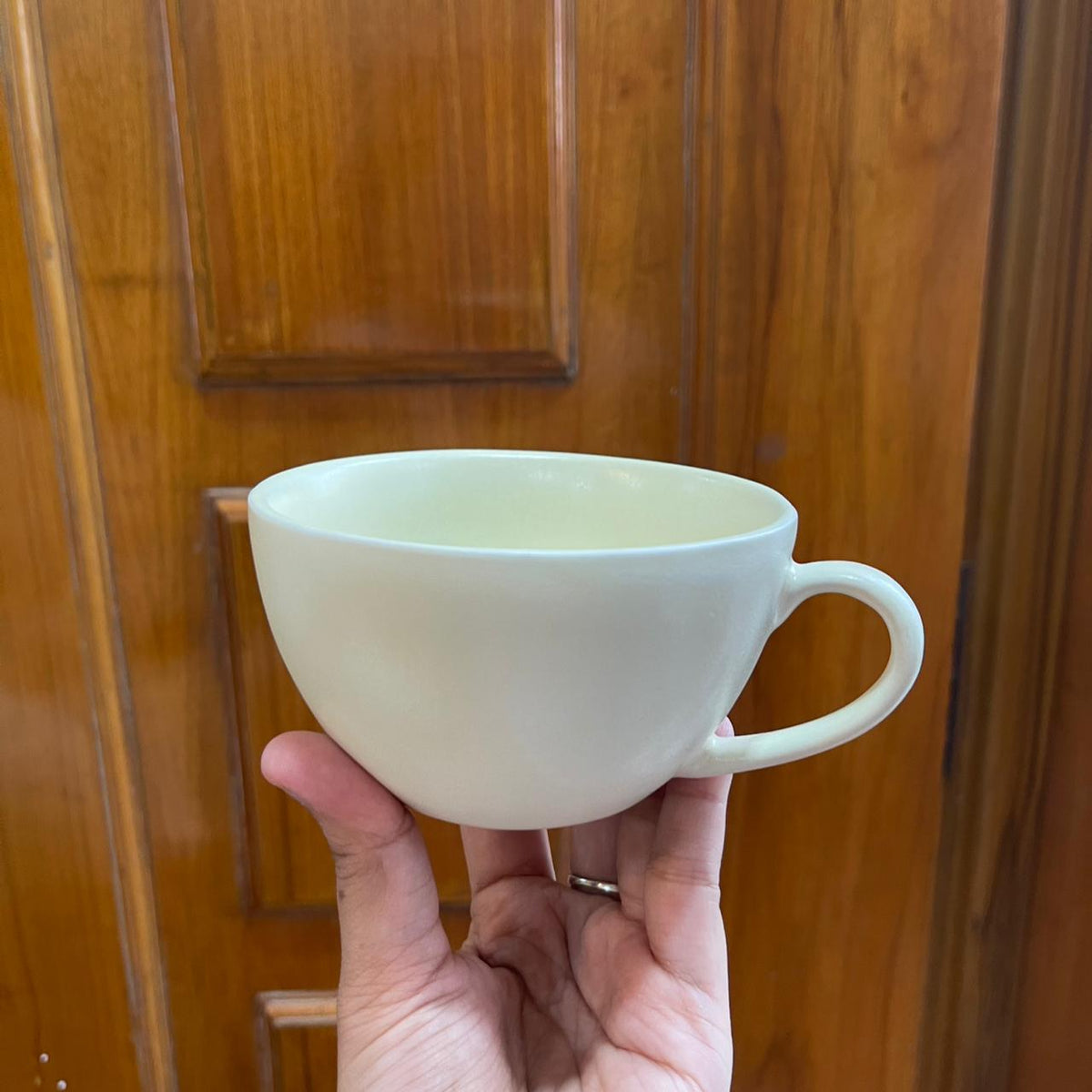 A hand holding a matte off-white ceramic cup in front of a warm brown wooden door.