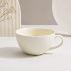 A minimalist ceramic cup with a rounded bowl and a curved handle, shown in a warm off-white or cream color. The cup is placed on a light surface with soft shadows, and decorative dried flowers and neutral background elements are visible.