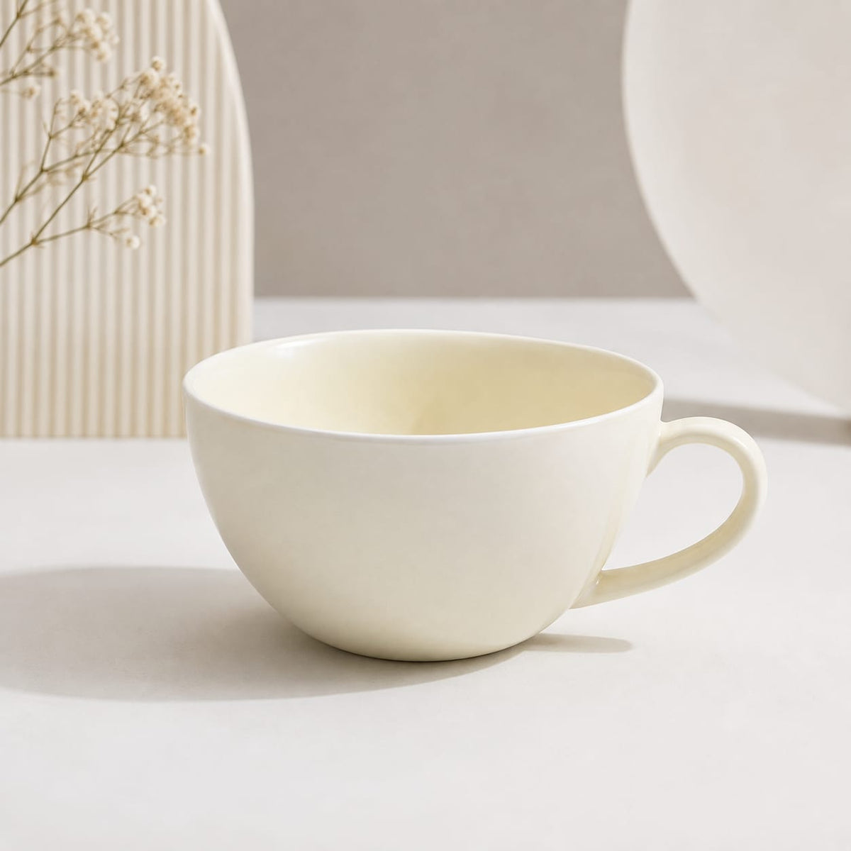 A minimalist ceramic cup with a rounded bowl and a curved handle, shown in a warm off-white or cream color. The cup is placed on a light surface with soft shadows, and decorative dried flowers and neutral background elements are visible.