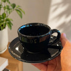 A glossy black ceramic coffee cup and saucer set being held by a hand, with a blurred potted plant in the background.