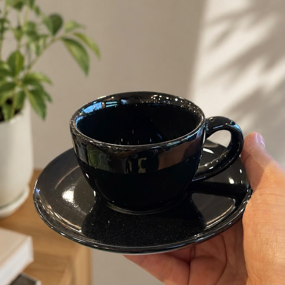 A glossy black ceramic coffee cup and saucer set being held by a hand, with a blurred potted plant in the background.